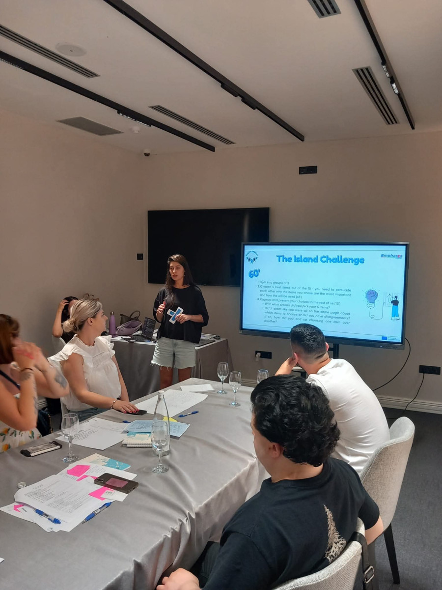 A  trainer is leading a workshop session, presenting content on a screen while participants sit around a table with notes and materials in a meeting room.