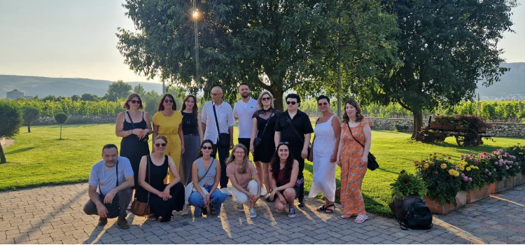 A group of training participants are posing outdoors in a well-maintained green area, with trees and vineyards in the background.