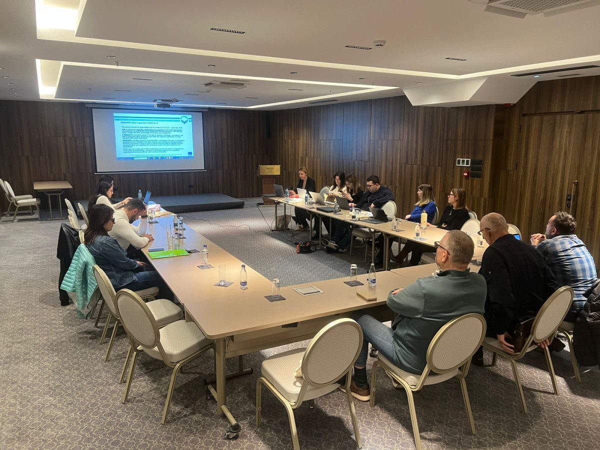 Project partners are seated around tables in an indoor meeting room during a meeting with a presentation on screen.