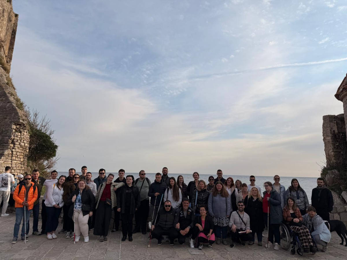The entire team of participants, volunteers, and leaders of national groups taking part in the youth training is standing for a photo on an outdoor stone pavement in the center of the coastal city of Budva. Old stone walls surround the group on the sides, while above them is a light blue cloudy sky. The atmosphere is cheerful, and the scene is spacious and inclusive.