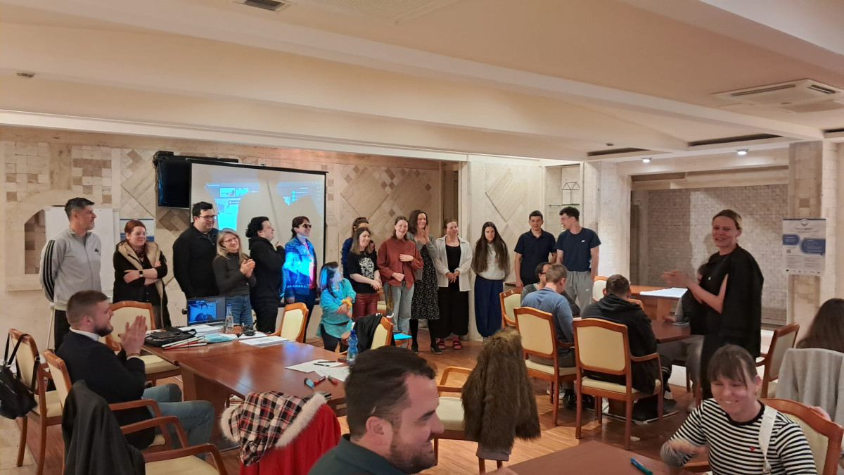 The volunteers and leaders of the national groups in the project are standing in a semicircle in a conference room, laughing and applauding. The other participants are sitting around the tables and are also applauding. In the foreground stands the leader of the entire project, a woman who is smiling and applauding as she thanks her team for their help in carrying out the whole training. The room is warmly lit.